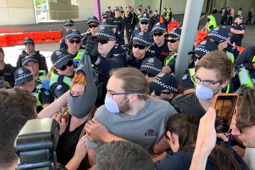 A crush of cops pressing in behind a line of link-armed resisters, the leftmost of whom has eyes closed in a grimace, orange hair exposed, balaclava pulled tight and up from face by the gloved fist of a cop close behind