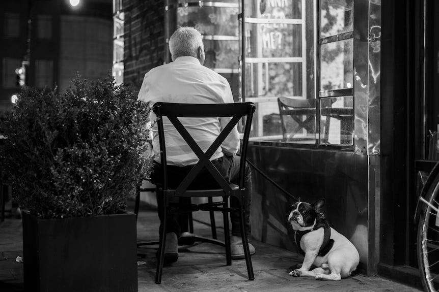 Senior man sat outside restaurant at table with his small dog beside him