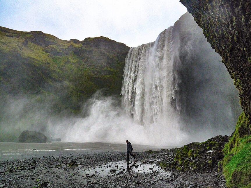 Man walking along water’s edge with tall waterfall behind