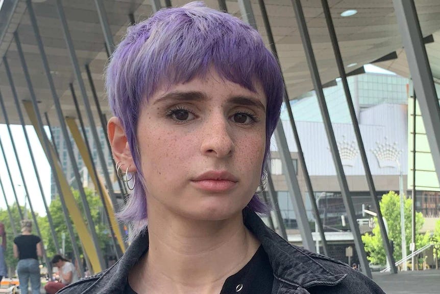 A young woman in close-up, dark eyes determined and a steady expression on her small, freckled face, with pastel purple hair that skims her collar and several silver hoops hanging from each ear