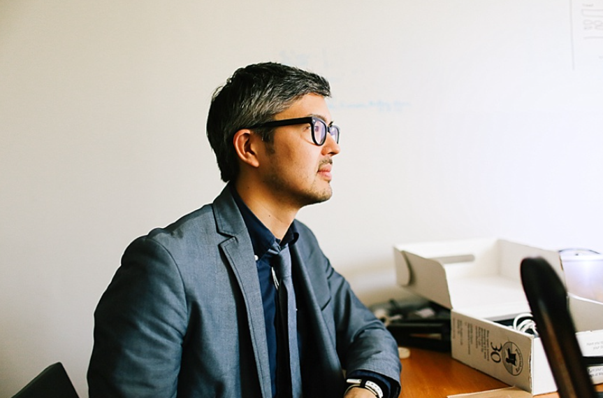 Black and grey haired mixed Korean-American person in a blue-grey blazer and loose tie over and black-rimmed glasses looking across a desk towards the right. Cardboard boxes and things cover the desk in the dimly lit room with white walls. Photo by Dakota Lenox Photography.