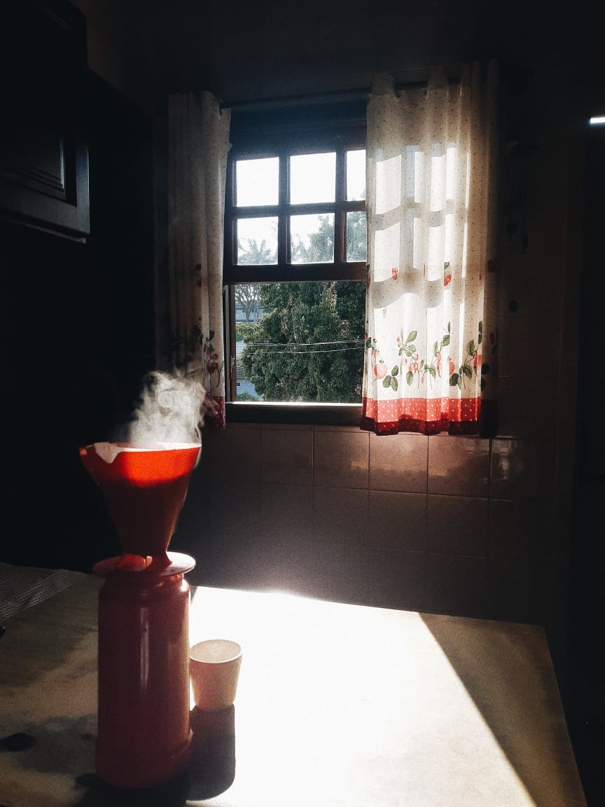 Uma térmica com filtro passando café sobre a mesa da cozinha posicionada em frente a janela.