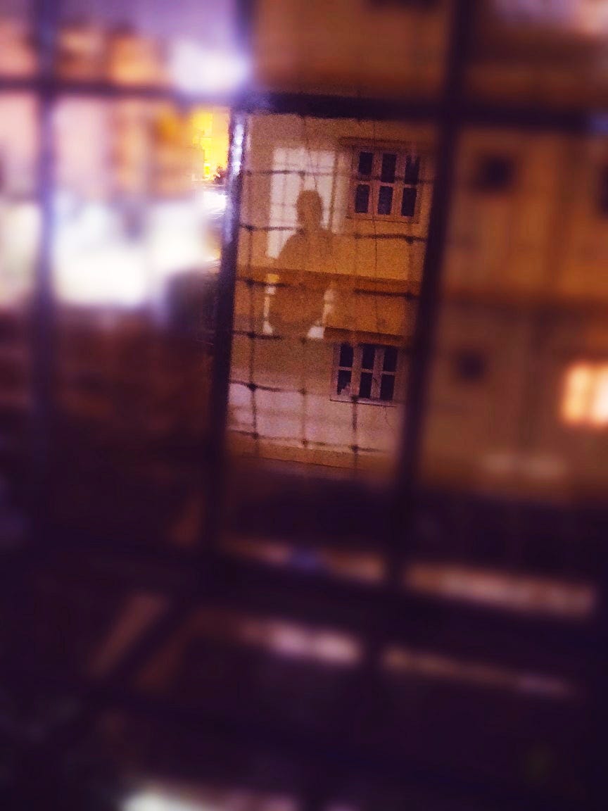 In the image, there is a girl standing on a balcony. Her shadow is cast upon the opposite wall. The buildings around are illuminated by bright city lights, which create a blurred, hazy effect in the picture.