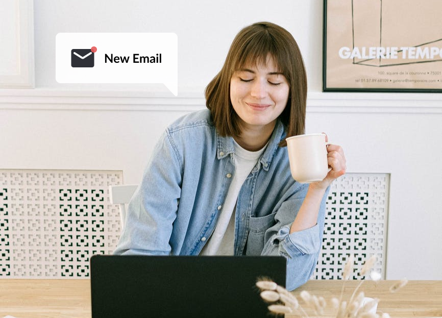 Woman sitting at table in front of laptop while holding a coffee mug and smiling at a “New Email” notification