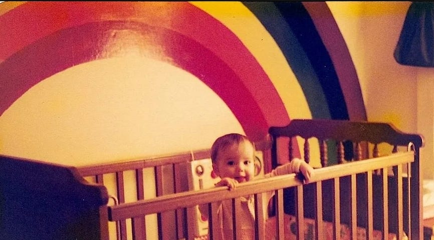 Baby me, Jenny Lane, standing in my wooden crib, hands holding the edge, painted on the wall of my room is a purple first rainbow