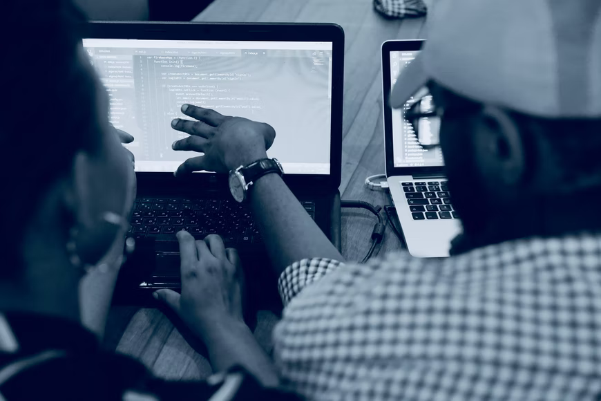 Foto em preto e branco e duas pessoas olham para tela de um computador. Uma delas aponta para a tela