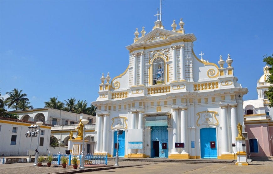 The Church of Our Lady of Immaculate Conception in Goa, India, featuring a white and gold Baroque-style façade with blue doors. A statue of the Virgin Mary is placed above the entrance, and two golden statues stand in the courtyard. The bright exterior contrasts with a clear blue sky.