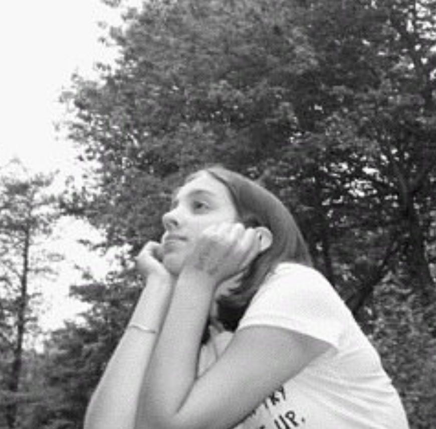 A young white female teenager sits with her head resting on her hands. The photo is in black and white and there are trees in the background. There is writing on the girl’s hand, but it is impossible to make out what it is. She is wearing a silver bracelet and a light-colored T-shirt.