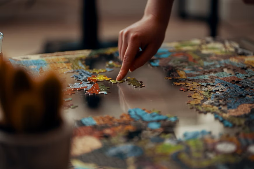 A hand of a human is pointed down, moving a puzzle piece into the location that is necessary for puzzle they are putting together upon a glass table surface.