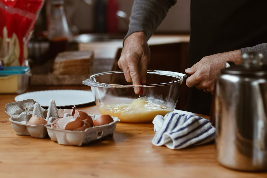 A person is shown with just their two hands. One hand is grasping a whisk and whisking the eggs in a clear glass bowl while the other hand is grasping the side of the bowl for stability. There is a small six space cardboard carton in front of the bowl with six open, cracked eggs.
