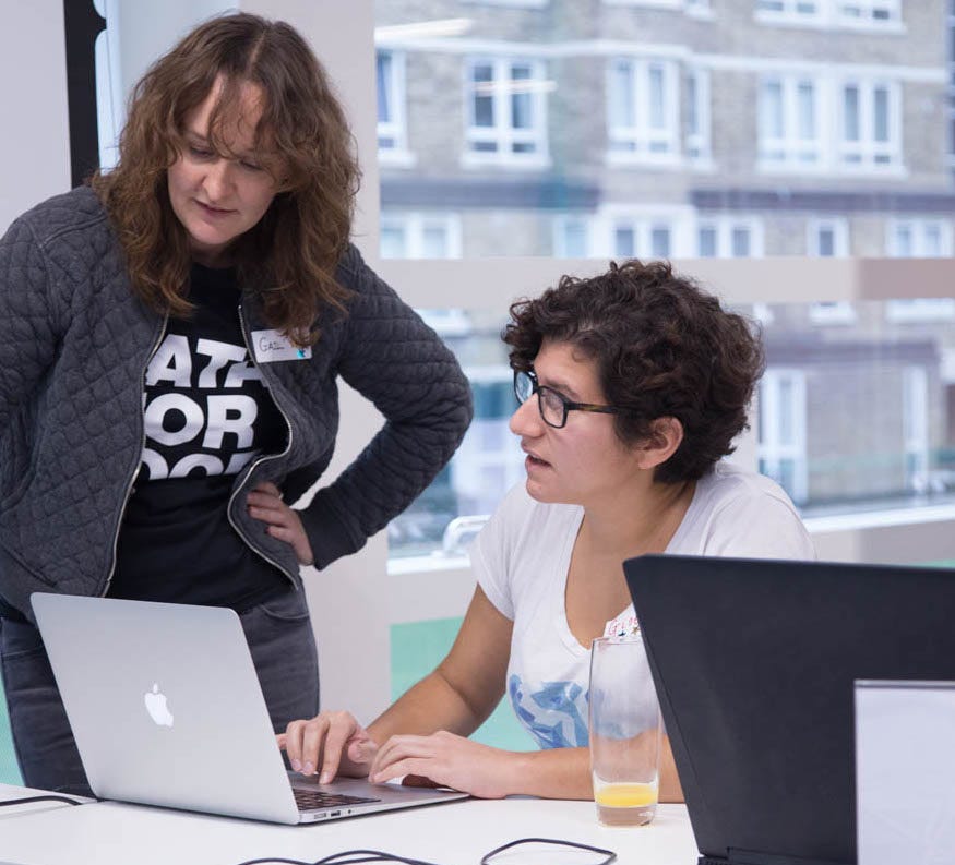 Giselle in three quarter profile, sat at a laptop with a window behind them, talking to Gail, who is stood to the left of the picture