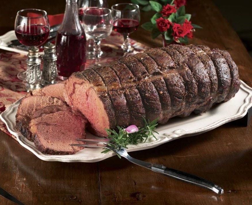 Medium-rare boneless Prime Rib on a white ornate platter, with glasses of wine and a small red bouquet on the table.