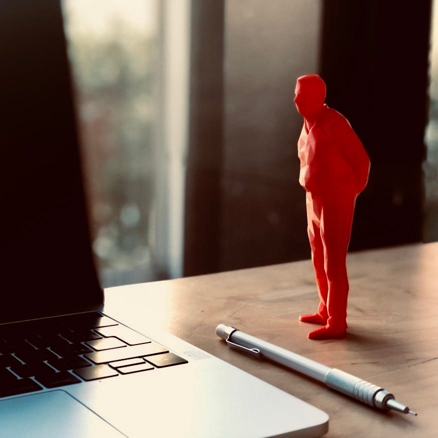 A 15 centimeter tall red plastic figurine of man with hands clasped on back “watching” your work without helping, placed on work desk by laptop.