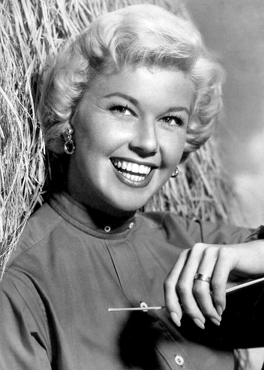 Doris Day, with blond curly hair, is leaning against a bale of hay, smiling and looking directly at the camera.