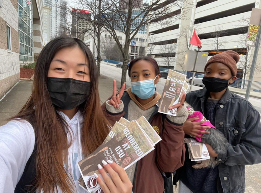 Three students wearing masks pose for a selfie, holding GlobeMed at GSU brochures.