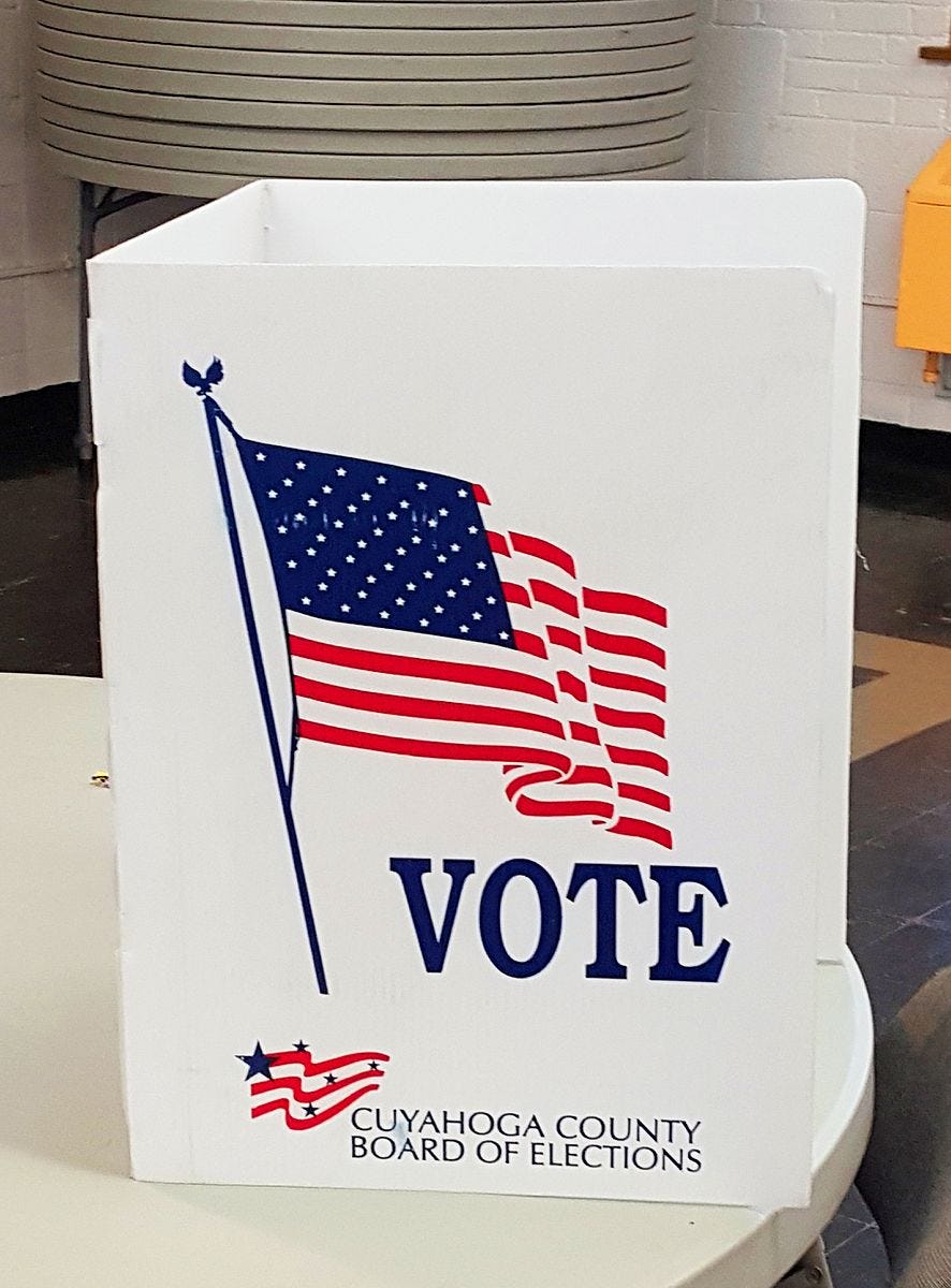 Photograph of a white voting booth with the US flag and the words “Vote” in blue on the side.