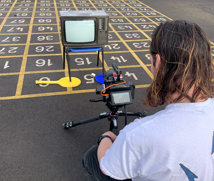 A man with long hair, his back to the camera holding a camera on a ground tripod, looking at a TV on top of a children’s number grid in a playground