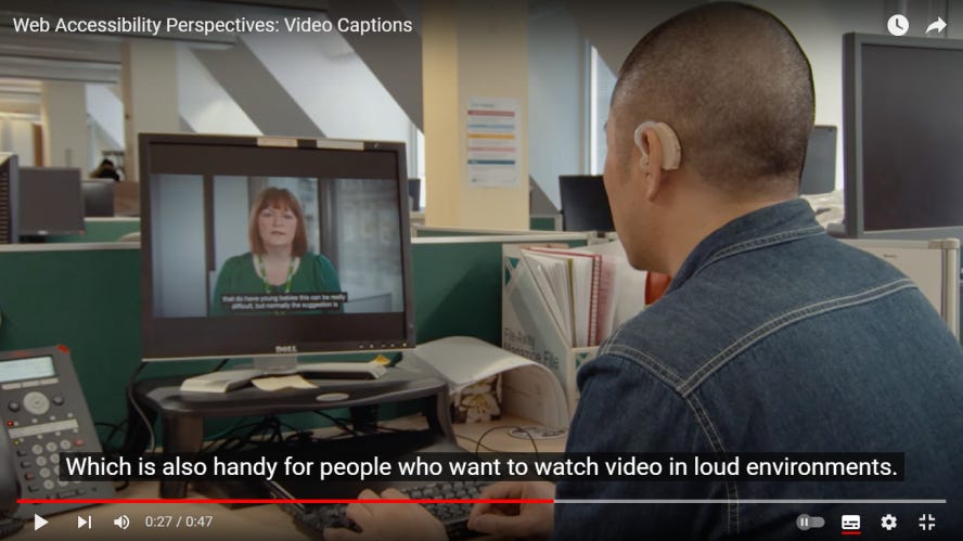 A YouTube video of person with a hearing aid sitting and watching a video on their computer. The video has closed captions enabled, which currently reads: “Which is also handy for people who want to watch video in loud environments.”