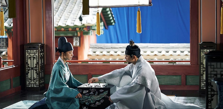 Two people dressed in ancient Korean (Joseon) clothes in a royal setting play a baduk (Go) game.