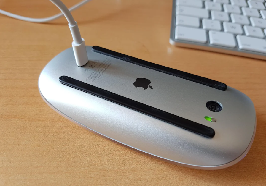 Apple Magic Mouse charging upside down on a wooden desk.