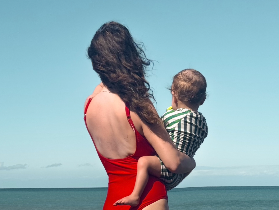 Alt text: photo of young mother holding child on her hip taken from the back as they both look out onto the sea in swimsuits