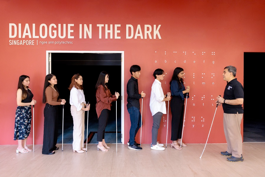 Seven people with canes standing in a single line, in front of them a blind tour guide. Writing on the wall: Dialogue in the dark, Singapore Ngee Ann Polytechnic. Braille alphabet on the wall.
