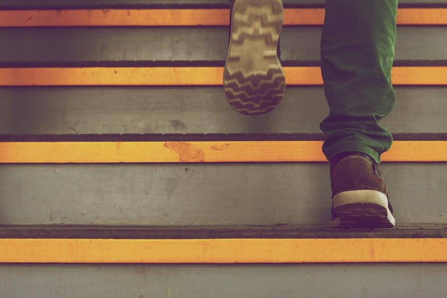 A close up of a person walking up stairs with just their shoes in view, wearing green pants. Each step has a yellow lip
