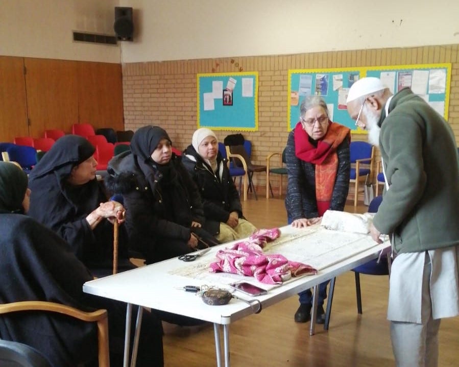 A group of women wearing headscarves are sat at a table listening to an older man and woman talking through the sewing materials in front of them.