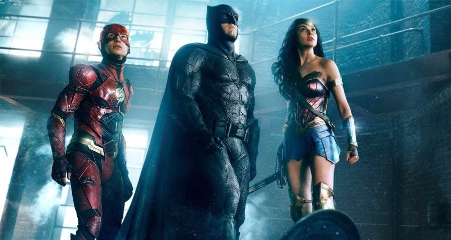 In front of a blue background, the Flash, Batman, and Wonder Woman from ‘Justice League’ (2017) are looking up.