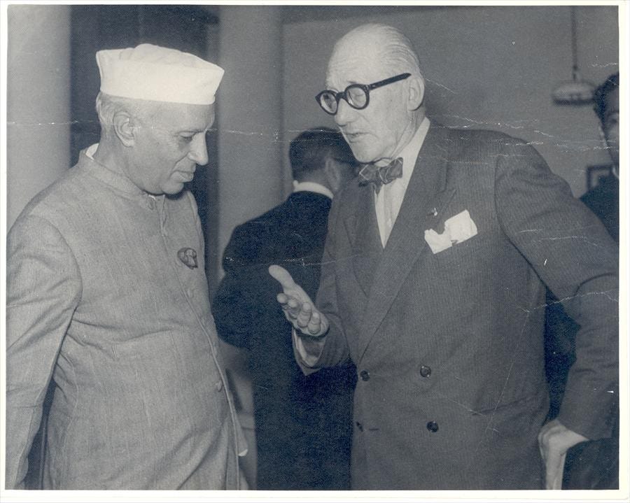 A black-and-white photograph of two men in conversation. One, wearing a Nehru jacket and Gandhi cap, listens attentively, while the other, in a suit with round glasses and a bow tie, gestures while speaking. The setting appears formal with other attendees in the background.