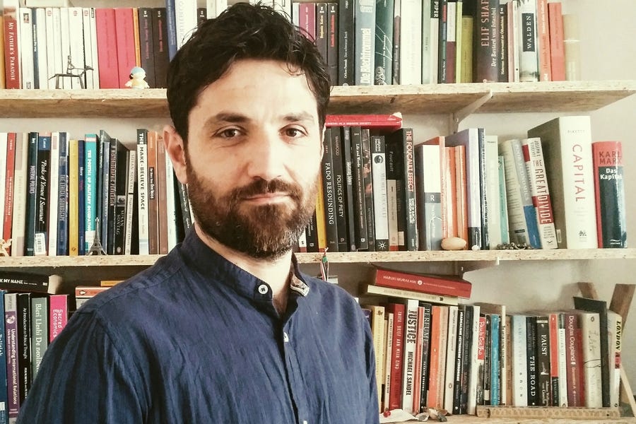Headshot photo of a man with a beard wearing a button-up shirt standing in front of a bookshelf.