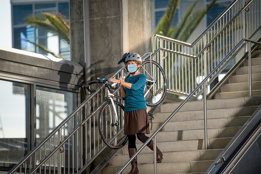 A person wearing a mask walks with their bike over their shoulder, down a long set of steps at a train station