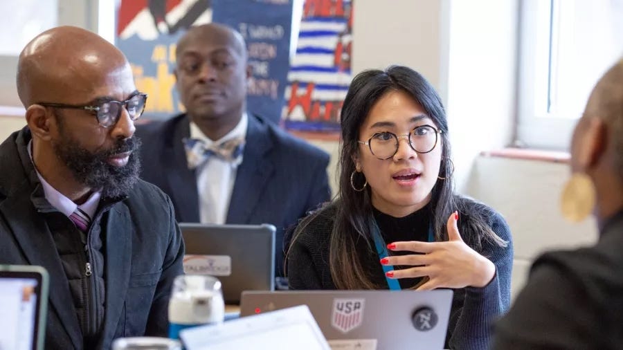 An Asian female teacher sitting next to a Black male teacher speaks to someone off-camera. Photo by Allison Shelley for EDUimages