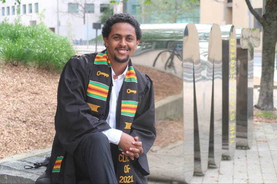 Man in graduation robes sitting in front of MIT sculpture.