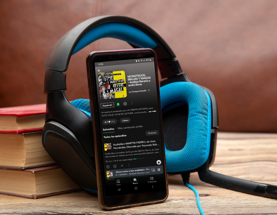 Auriculares celestes y libros con screenshot del podcast MONSTRUOS, BRUJAS Y MAGAS, en Spotify en donde se comparten audiolibros en español