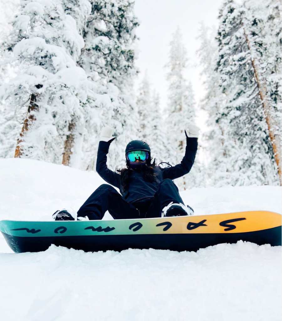 Snowboarder sitting on the mountain in powdery snow looking excited while holding up both arms. He is riding a Salomon snowboard.