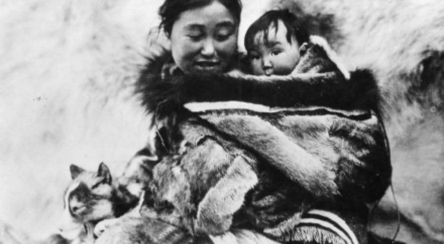 A young Inuk woman carrying her baby on her back, both swathed in animal fur.