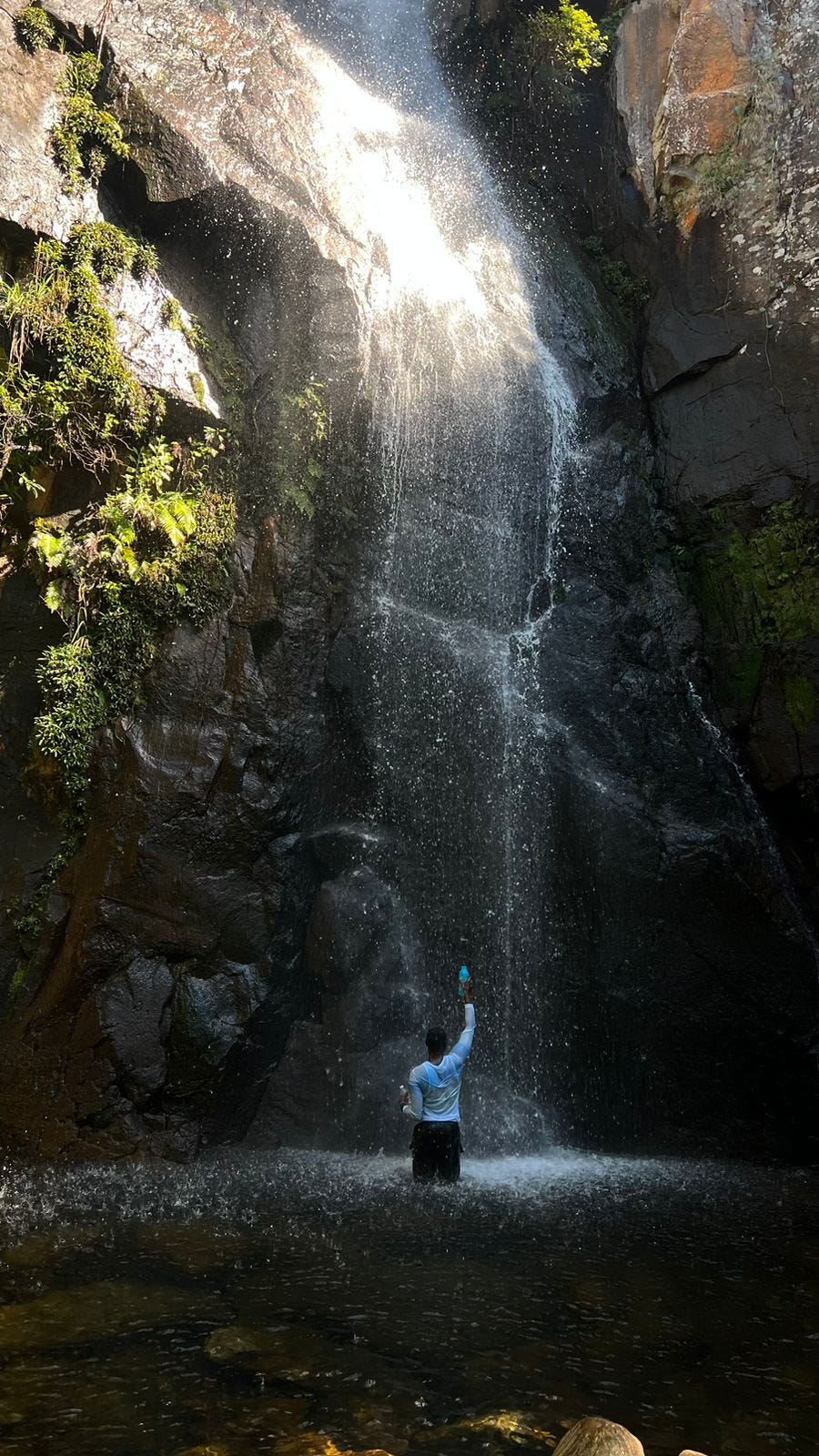 An African male holding a water bottle above his head standing at the base of a massive waterfall cascading over a rock cliff