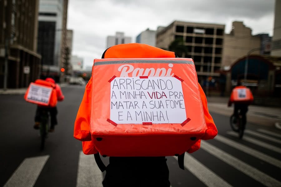 A photograph of an Anti-Fascist Deliverers riding his bicycle from the back. We can see a handwritten note glued on his backpack, partially covering the logo Rappi. The backpack has a very bright orange colour, contrasting with the grey shades of the city pavement, the buildings and the cloudy sky.