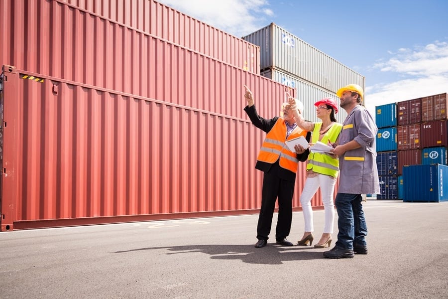 Three construction works wearing safety vests and helmets are holding clipboards and pointing to the shipping containers surrounding them.