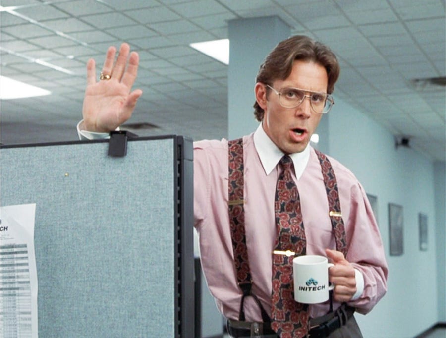 Early 1990s picture of a business executive in an appalling pink shirt with white collar and loud tie and braces combo. He’s holding a white mug with ‘Infotech’ written on it, leaning over a cubical partition with an expression that says he’s just about to ruin someone’s day.