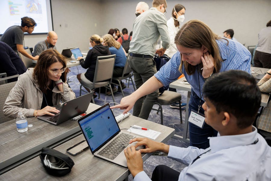 More than a dozen people sit around shared tables with laptops running App Inventor.