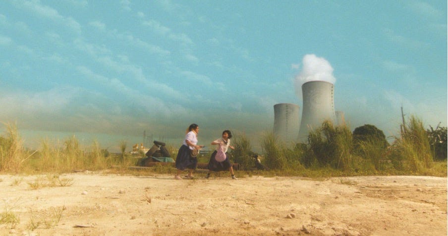 From a distance we see two high school girls engaged in a fistfight in a dusty field surrounded by thick yellowing grass. Behind them looms two gigantic nuclear cooling towers billowing thick clouds of white hot steam.