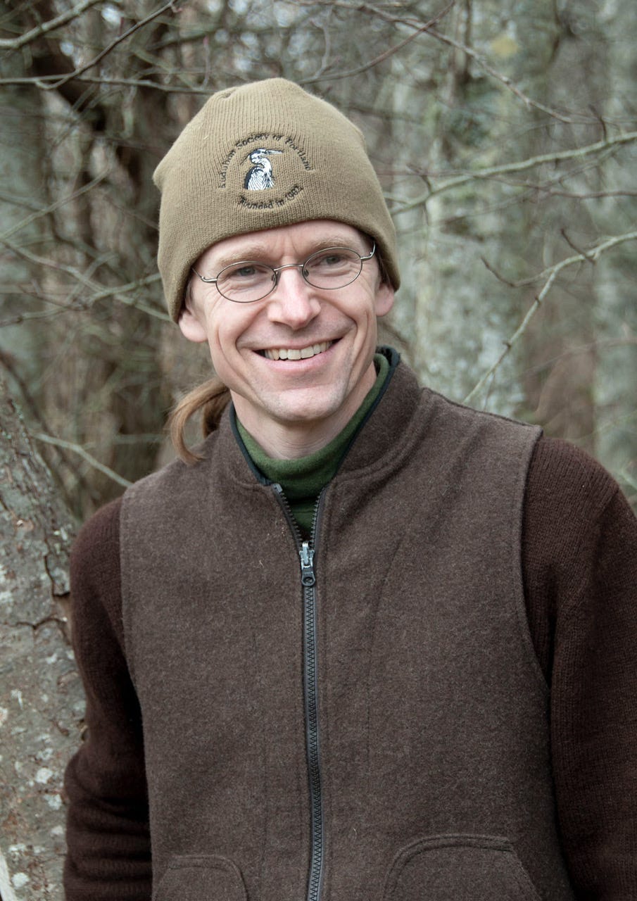 Close up shot of author Thor Hanson smiling, wearing a khaki hat, green turtleneck and brown jacket, in a forest.