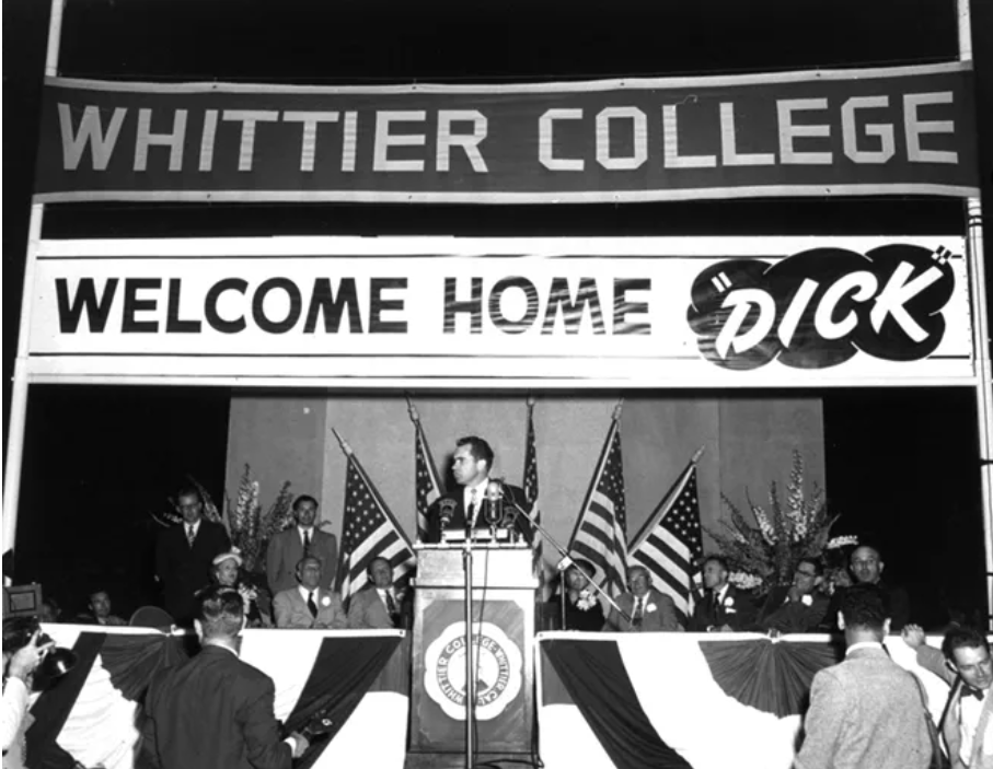 Black and white photo of Richard Nixon standing on stage in front of a podium with banners behind him reading “Whittier College” and “Welcome Home ‘Dick’.”