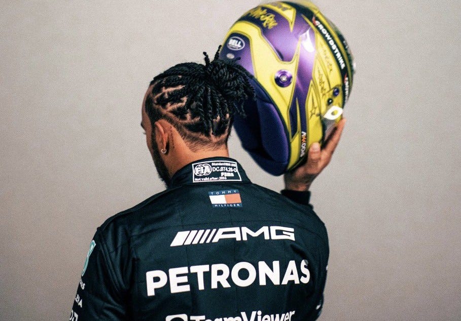 Lewis Hamilton wearing his race suit with his back turned, holding his helmet up to one side of his head.