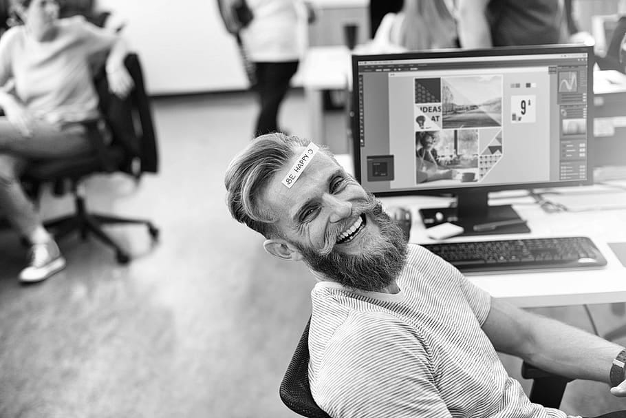 A man in an office with a post-it note on his forehead saying “be happy”.