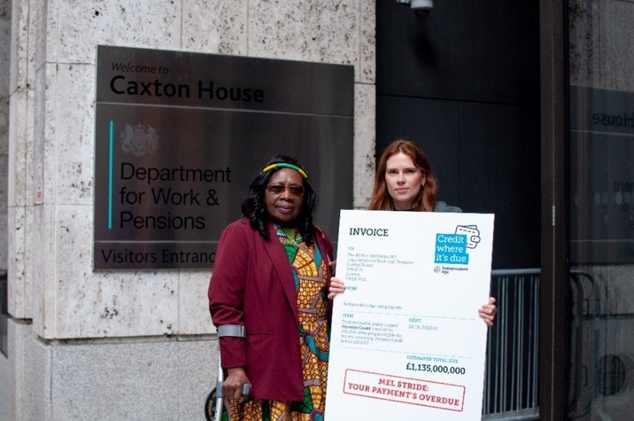 Photo of an older woman wearing a colourful dress and using a crutch, stood next to a younger woman who holds a large invoice prop telling Mel Stride that the Government’s Pension Credit payments are overdue.
