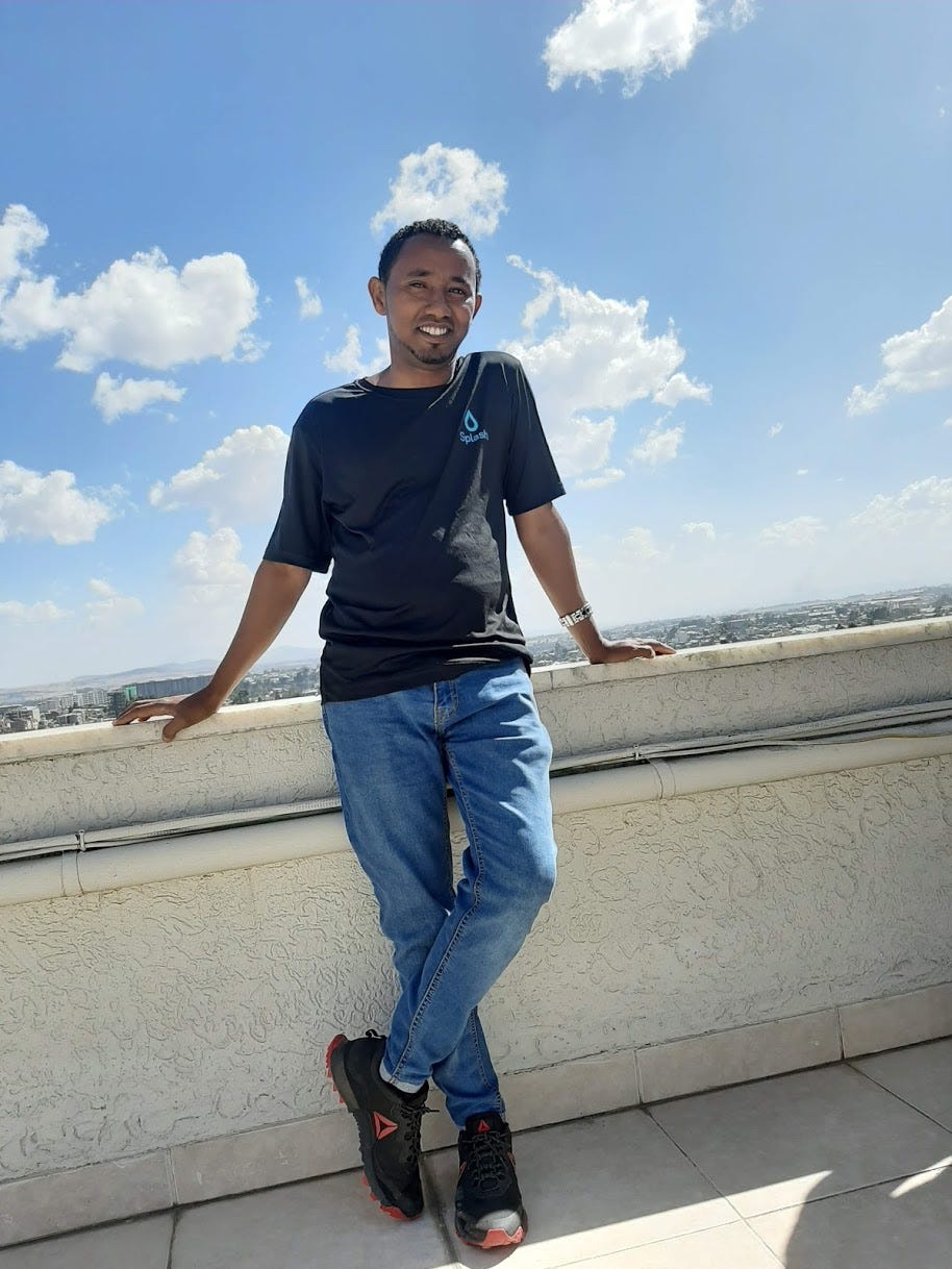 Splash staffer Nasser Ferej stands smiling on a rooftop in a black shirt and jeans, with a beautiful blue sky and picturesque clouds in the background.