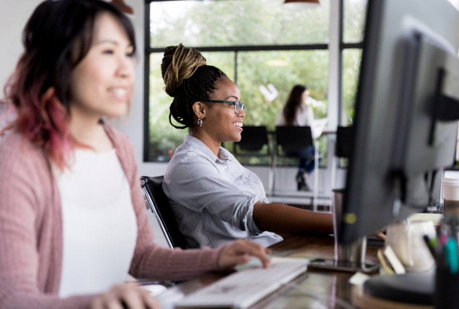 An Asian woman with pink hair and a Black woman with her hair up in a bun sitting in from of their computer screens, smiling. Credit: Canva